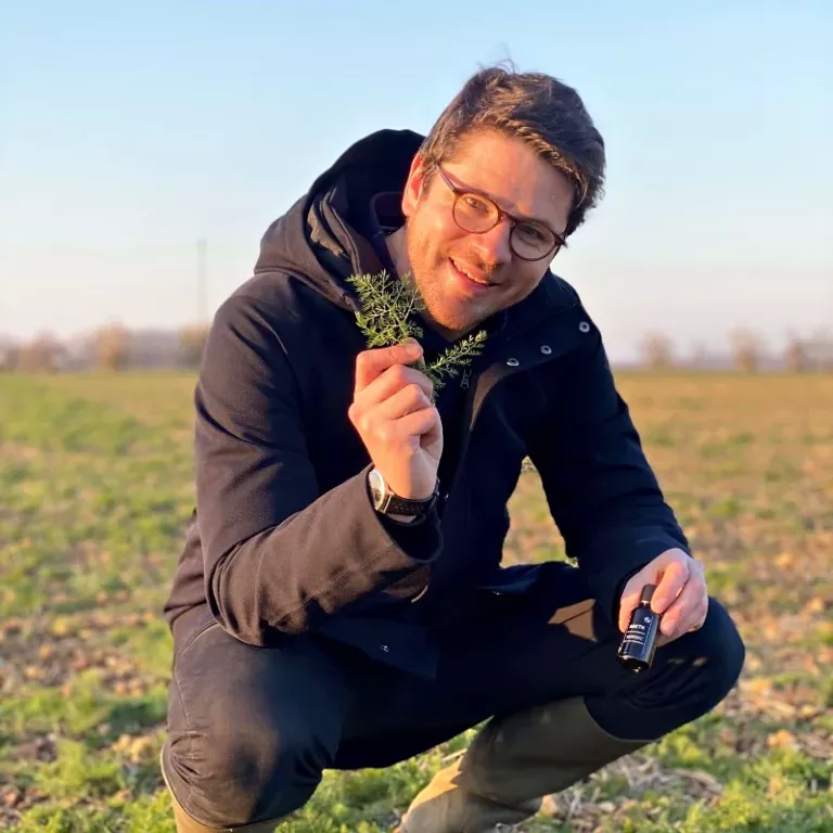 Valentin Raoul, producteur, accroupi dans un champ de Foucrainville avec un brin d’aneth et un flacon d’huile essentielle d’aneth