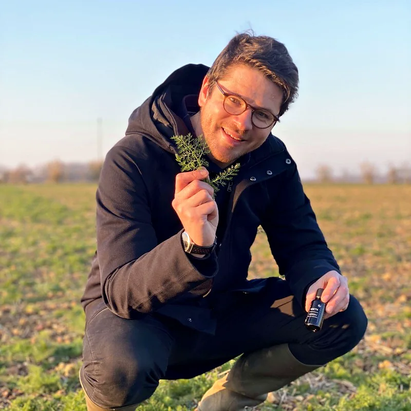 Valentin Raoul, producteur, accroupi dans un champ de Foucrainville avec un brin d’aneth et un flacon d’huile essentielle d’aneth