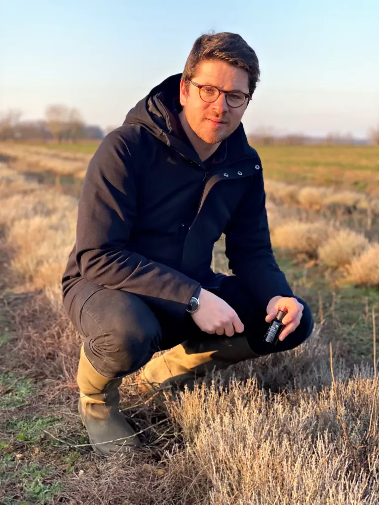 Valentin Raoul accroupi entre les rangs de plantes aromatiques, en hiver, un flacon à la main